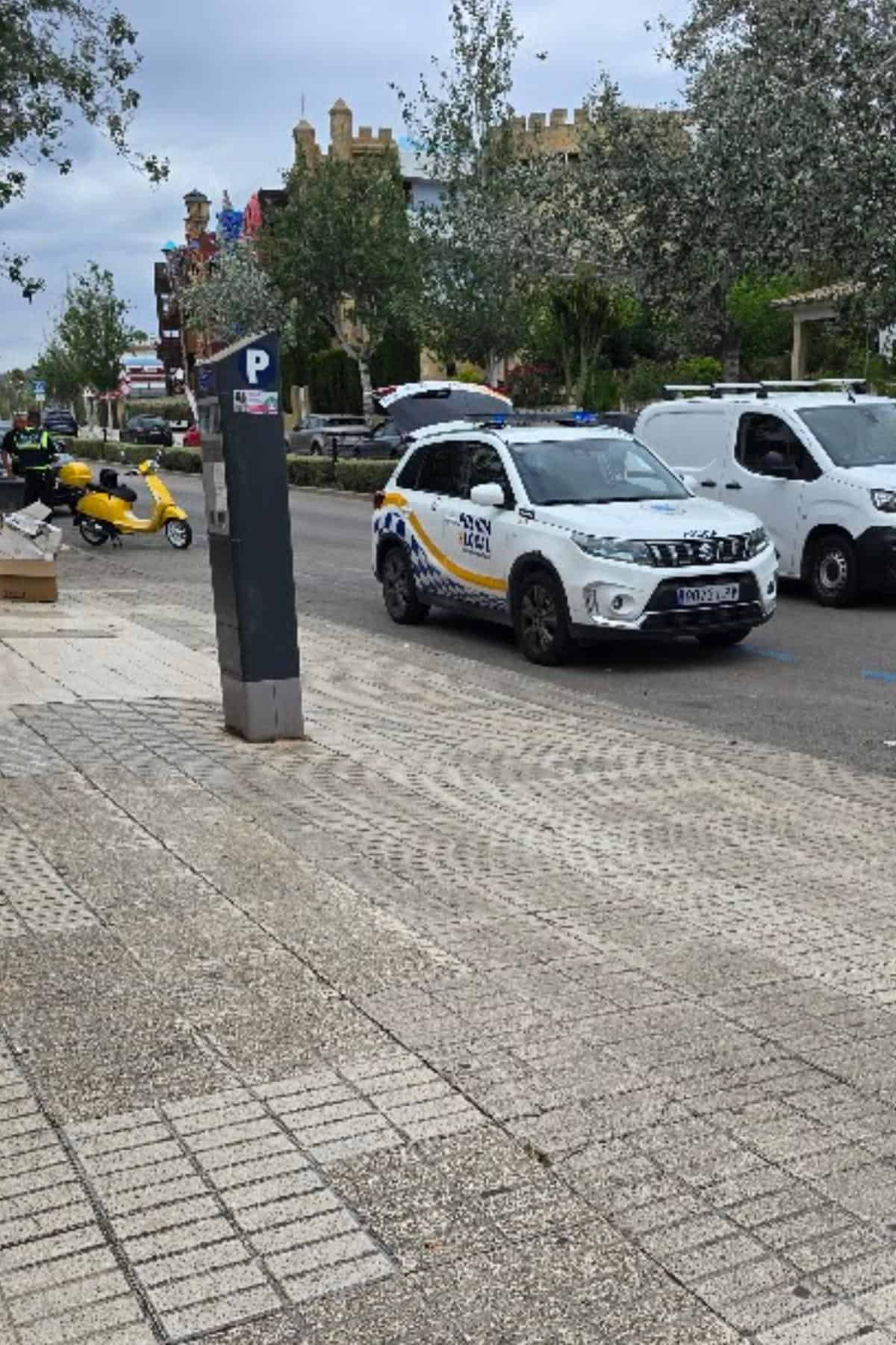 A city street scene perfect for a mallorca driving guide: a parked traffic police suv, yellow scooter, and white van. Pedestrians stroll beneath trees and an overcast sky, with a parking meter in the foreground—a glimpse of driving in mallorca.