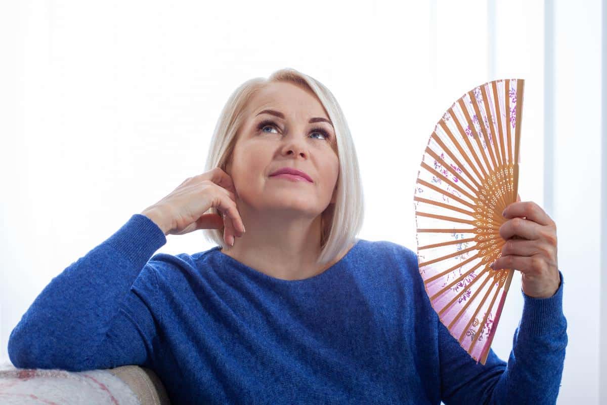 A middle-aged woman with short blonde hair, wearing a blue sweater, holds a decorative hand fan and looks thoughtfully upward while sitting indoors, perhaps reflecting on perimenopause.