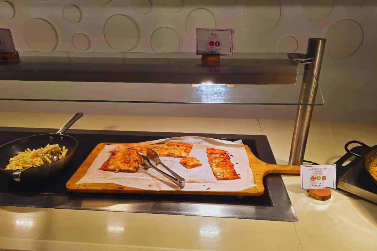 A buffet setup at Bahia del Sol in Santa Ponsa features rectangular pizza slices on a wooden board lined with parchment paper, metal tongs, a pan of fries to the left, and small signs labeling the all-inclusive food options.