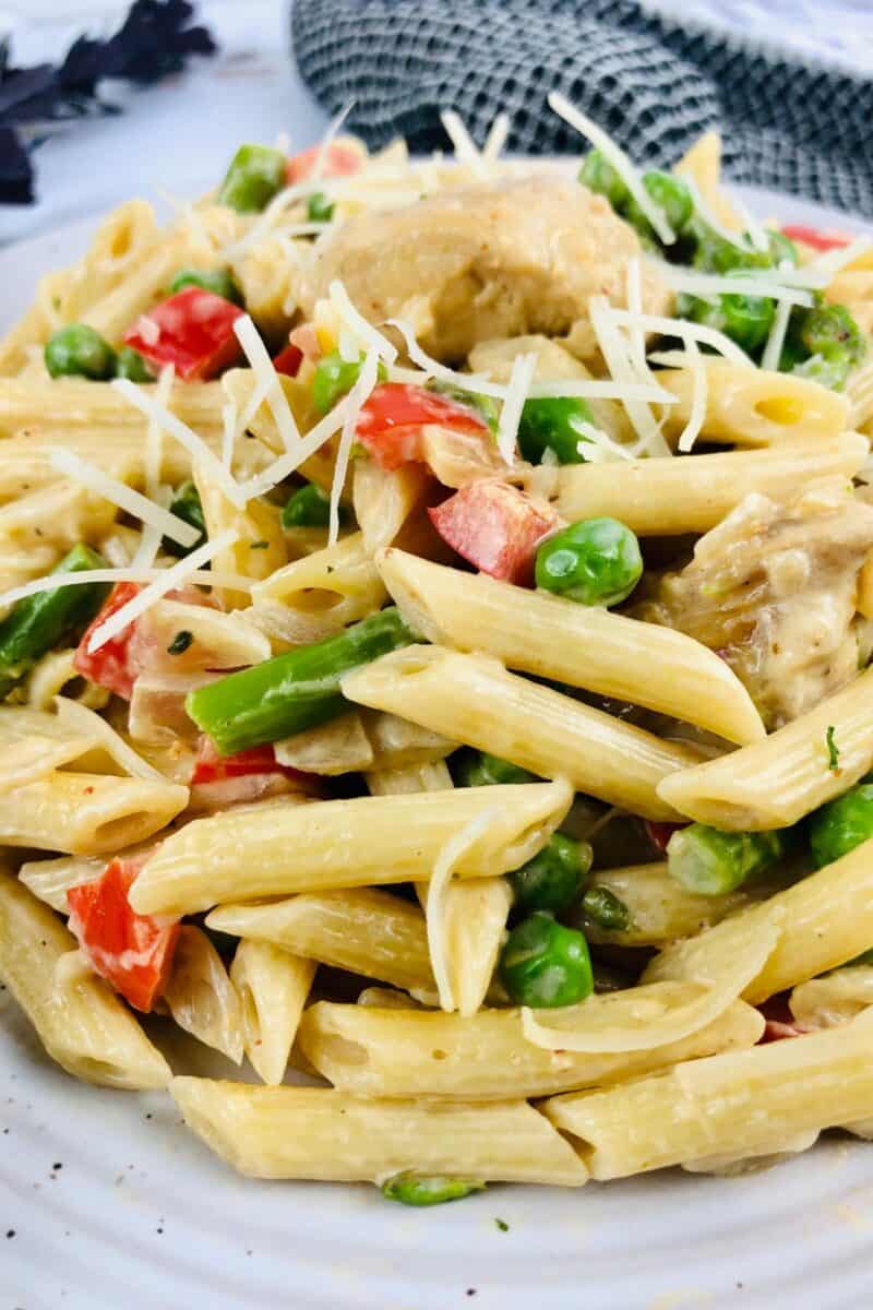 A close-up of creamy Chipotle Chicken Pasta mixed with green peas, asparagus, and red bell peppers, topped with shredded cheese, served on a white plate.
