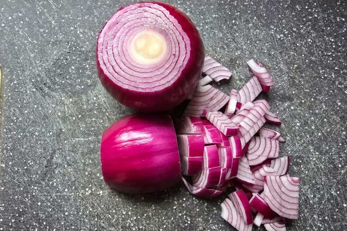 Two red onions sit on a dark speckled cutting board; one is whole with the top cut off, and the other is partly chopped into cubes and slices, ready to be used in your favorite chicken bhuna recipe.