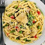 A white plate of Healthy Chipotle Chicken Pasta with chunks of chicken, green peas, red bell peppers, asparagus, and grated cheese is served with a fork and knife on a rustic white table with a black patterned napkin. Tastes amazing!.