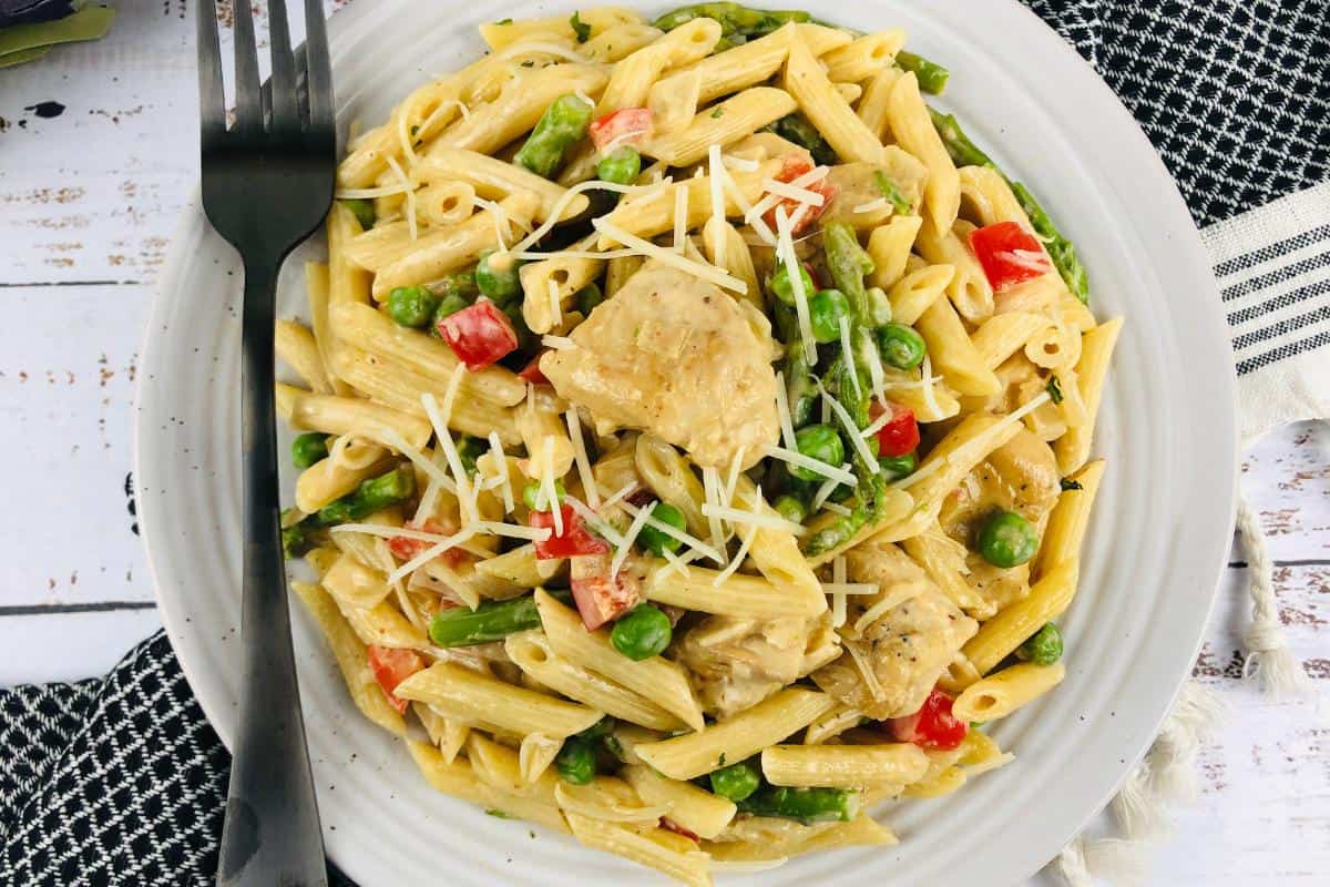 A white plate of Healthy Chipotle Chicken Pasta with chunks of chicken, green peas, red bell peppers, asparagus, and grated cheese is served with a fork and knife on a rustic white table with a black patterned napkin. Tastes amazing!.