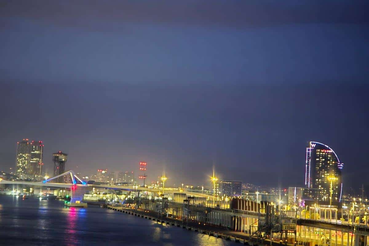 Nighttime cityscape featuring illuminated buildings and a waterfront with piers, evoking the allure of an overnight journey. Colorful lights reflect on the water, and a tall, curved building stands out on the right against the dark sky.