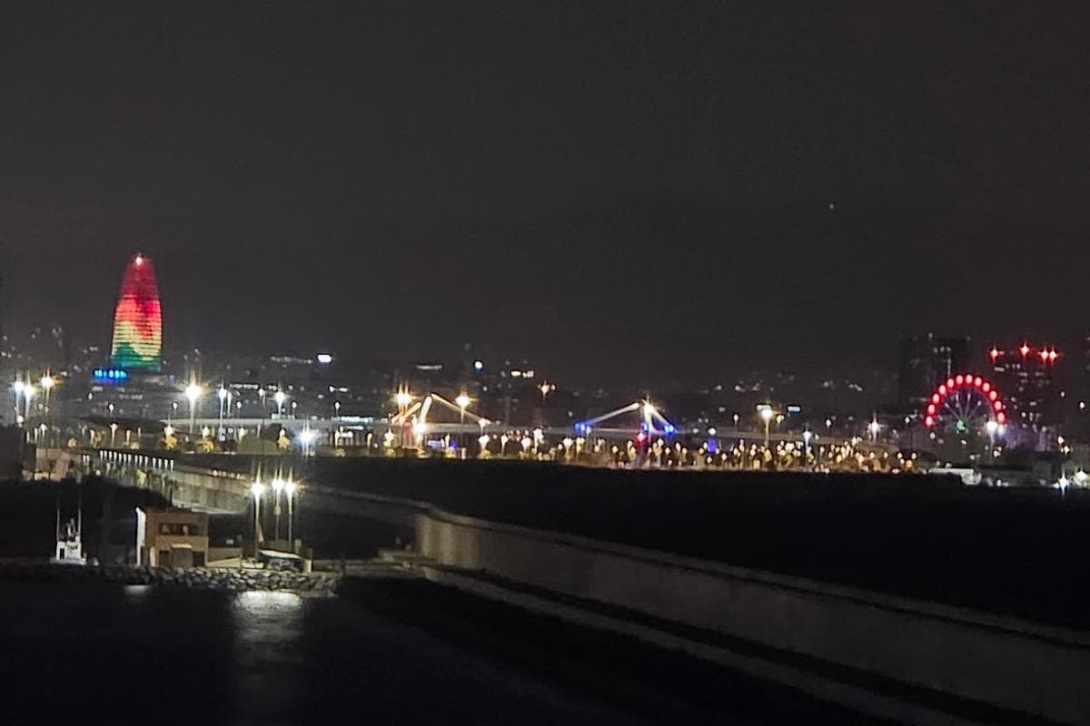 Night cityscape featuring a colorfully lit sail-shaped building on the left and the red GNV Ferries Ferris wheel on the right, evoking the spirit of an overnight journey from Barcelona to Palma, with streetlights brightening roads below.