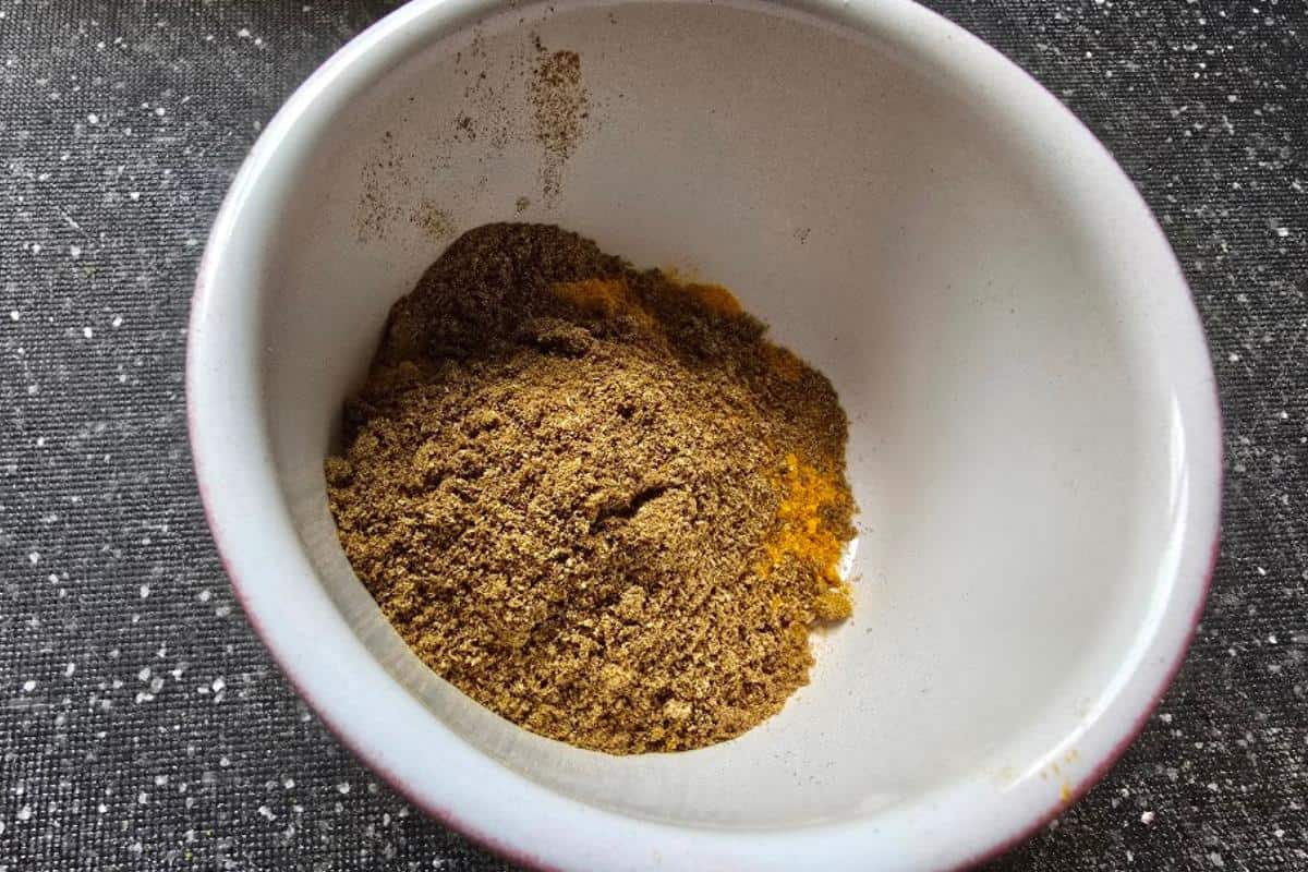 A small white bowl containing a pile of ground spices, mostly brown with a hint of yellow, sits on a dark, speckled countertop—perfect for adding depth to your chicken bhuna recipe.