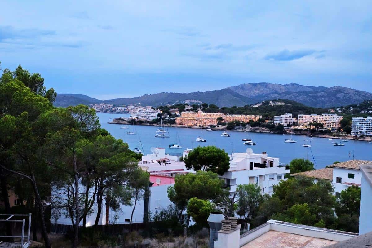 Enjoy a stunning view of Santa Ponsa, where white and pastel-colored buildings overlook sailboats anchored in the calm Bahia del Sol, framed by green trees and rolling hills under a cloudy blue sky—perfect for an all-inclusive getaway.