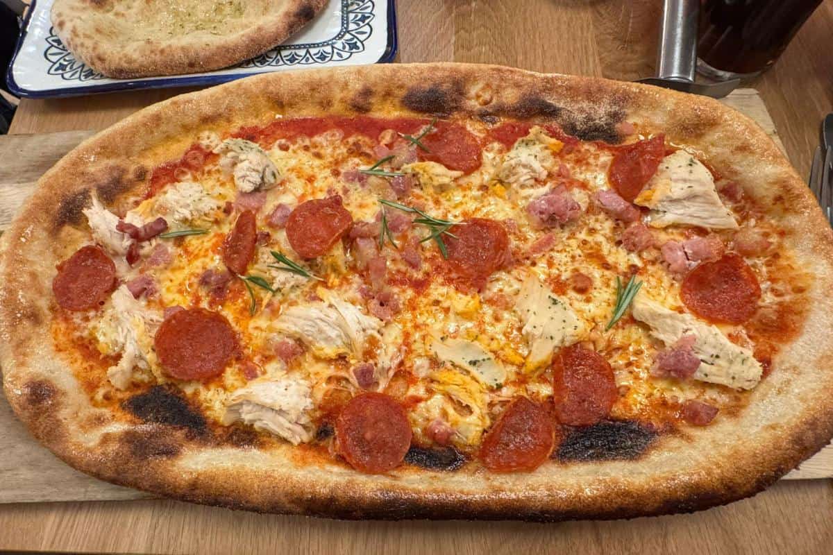 A rectangular pizza from Prezzo Italian Nottingham, topped with pepperoni, chicken, bacon bits, herbs, and melted cheese on a golden brown crust with charred spots, served on a wooden board—perfect for your next Nottingham restaurant visit.