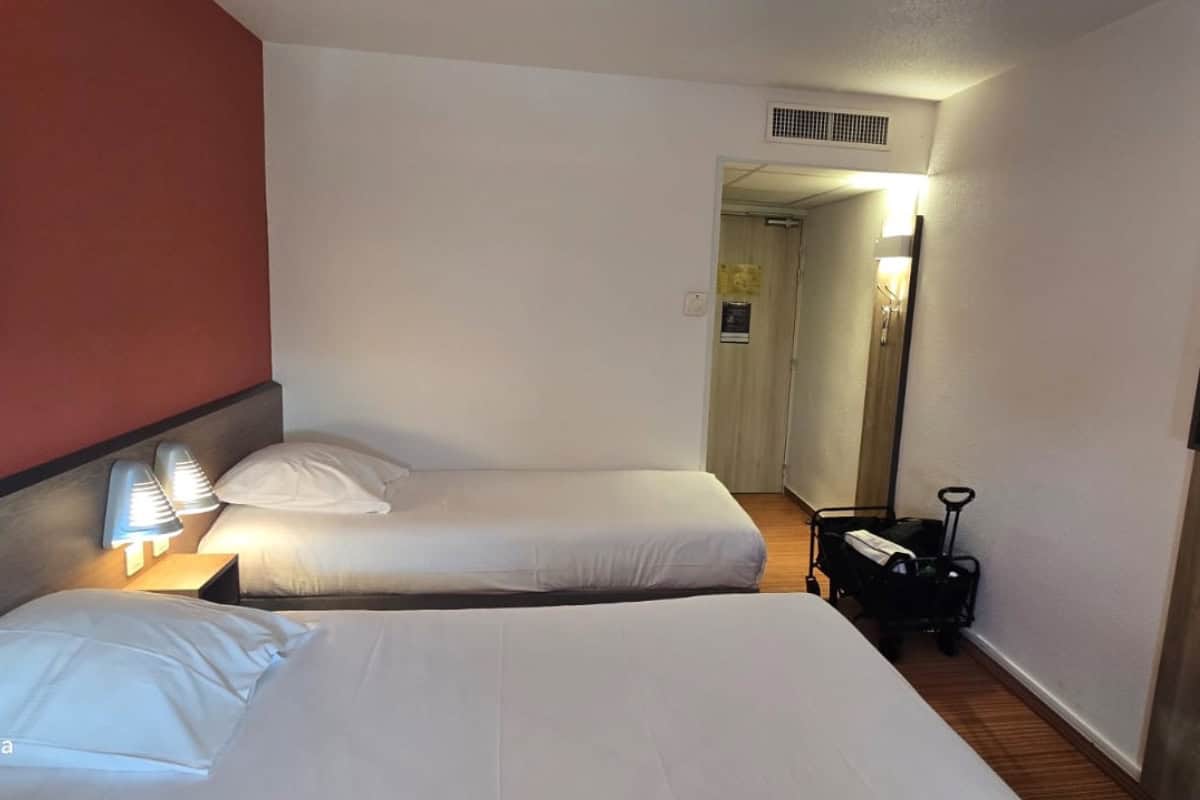 Hotel room with two single beds, a bedside table with two wall lamps, a luggage trolley near the door, and white and red walls.