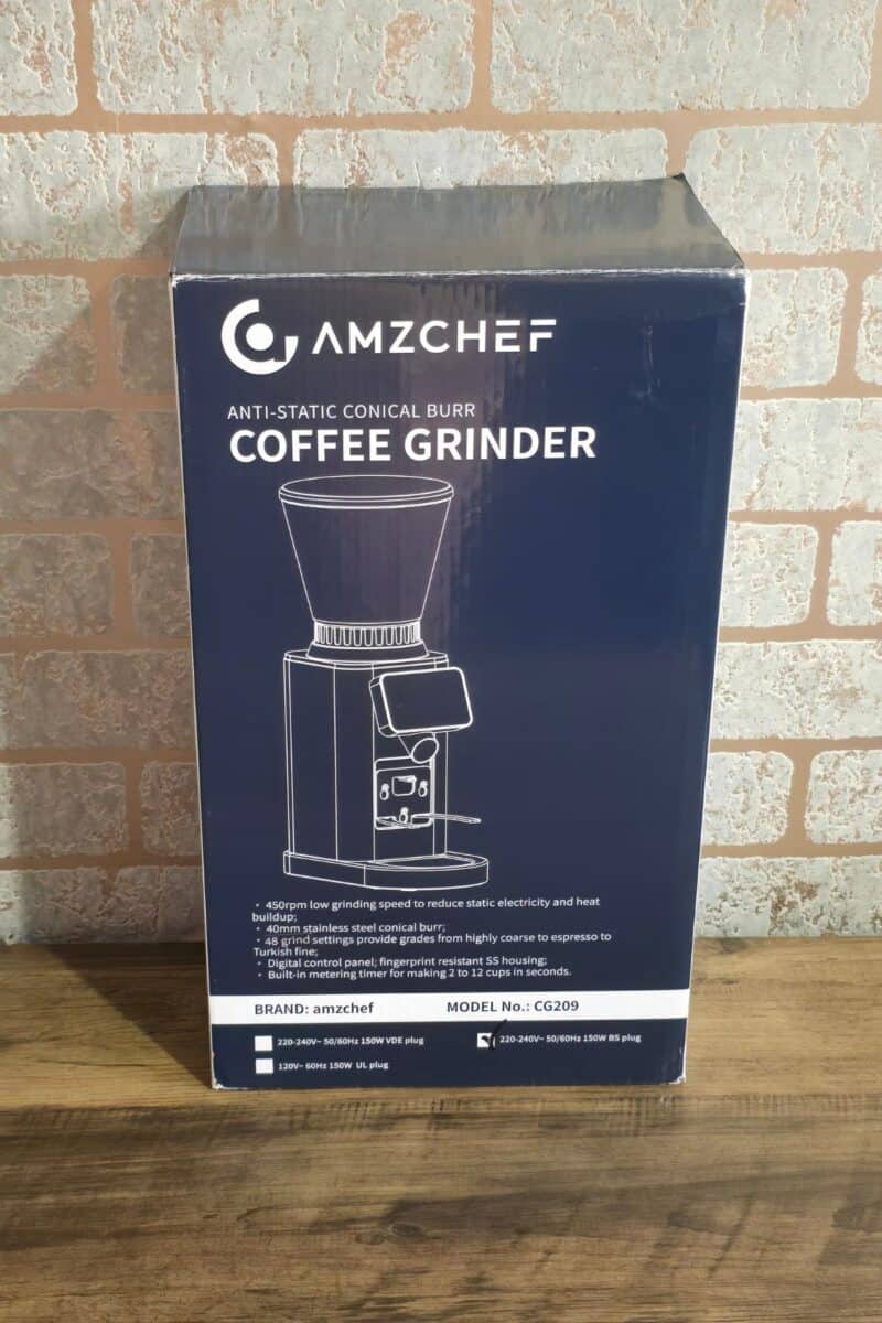 A boxed AMZCHEF anti-static conical burr coffee grinder is displayed on a wooden surface against a brick wall, featuring product highlights and a line drawing, promising perfect results with freshly ground beans.