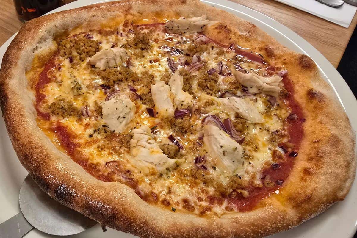 A close-up of a baked pizza topped with chicken, red onions, mozzarella, tomato sauce, and herb seasoning on a thick, golden-brown crust. Served on a white plate, this dish is a highlight at Prezzo Italian Nottingham.