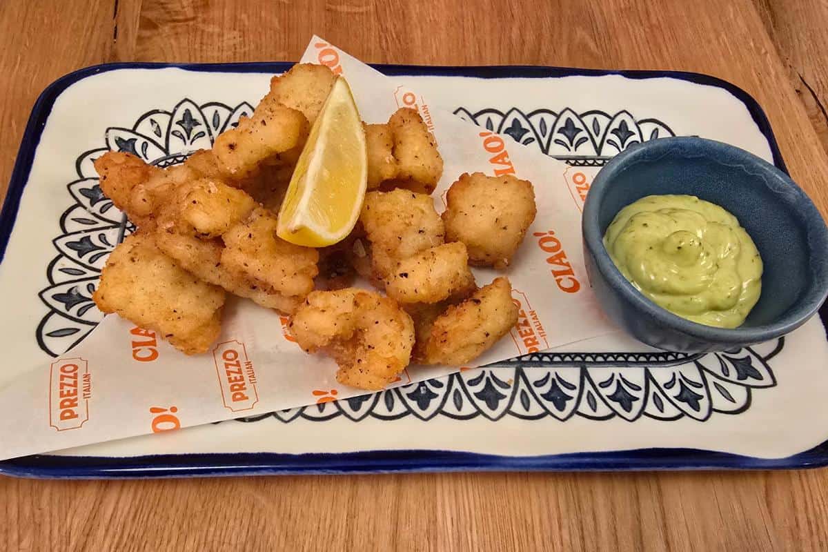 A plate of golden fried calamari pieces with a lemon wedge, served on decorative paper and a rectangular dish at Prezzo Italian Nottingham, alongside a small bowl of creamy green dipping sauce—perfect for your next review.