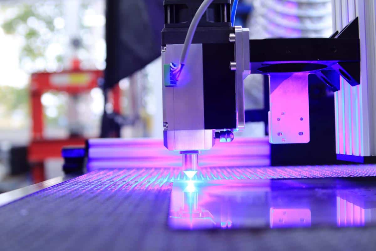 A close-up of a CNC laser cutting machine in operation, emitting a bright blue light as it precisely cuts or engraves a flat sheet for intricate design work on a worktable in a modern industrial setting.