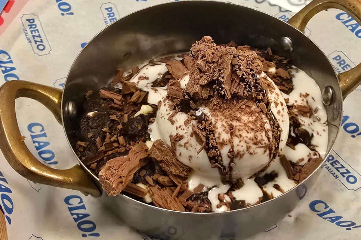 A metal bowl with handles filled with vanilla ice cream, chocolate sauce, and chocolate shavings, served on paper reading "CIAO!" and "PREZZO Italian" in blue at Prezzo Italian Nottingham. A sweet highlight in any Nottingham restaurant review.
