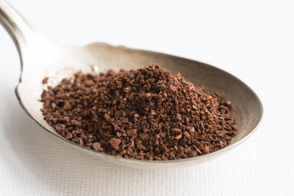 A close-up of a metal spoon holding a mound of coffee made from freshly ground beans, with a white textured surface in the background.