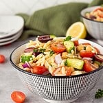 A bowl of healthy orzo salad with cherry tomatoes, cucumber, olives, chickpeas, red onion, and parsley sits on a gray surface. Another bowl, a lemon wedge, and scattered salad ingredients are in the background.