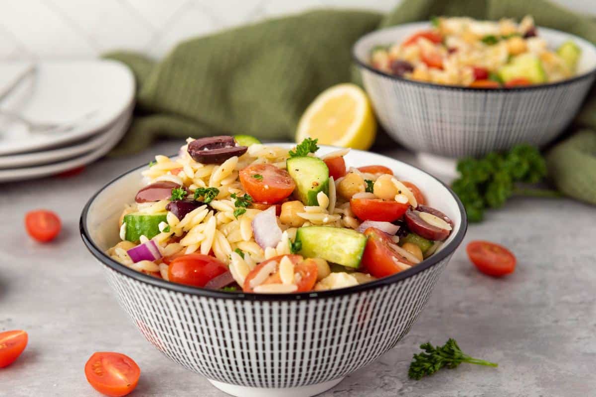 A bowl of healthy orzo salad with cherry tomatoes, cucumber, olives, chickpeas, red onion, and parsley sits on a gray surface. Another bowl, a lemon wedge, and scattered salad ingredients are in the background.