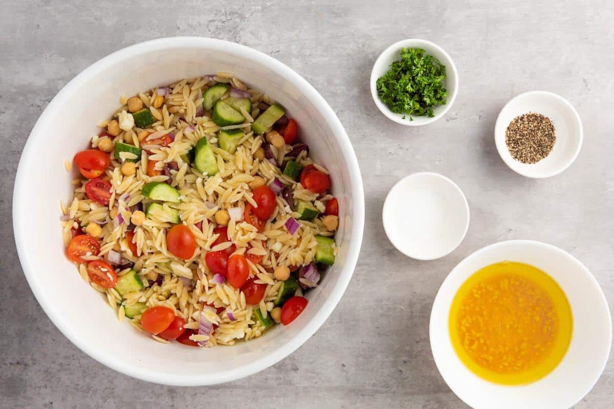 A white bowl filled with Healthy Orzo Salad—featuring cherry tomatoes, cucumbers, red onions, and chickpeas—sits on a gray surface. Nearby are small bowls of chopped herbs, pepper, salt, and a yellow vinaigrette for the ultimate healthy salad experience.
