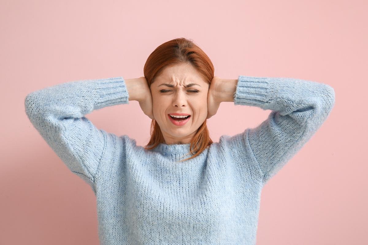 A woman in a light blue sweater stands against a pink background, covering her ears with her hands and grimacing, as if reacting to an unpleasant food noise.