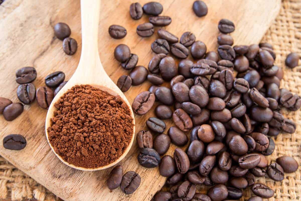 A wooden spoon filled with freshly ground beans rests on a wooden surface next to a pile of whole coffee beans. The background includes a woven burlap texture.