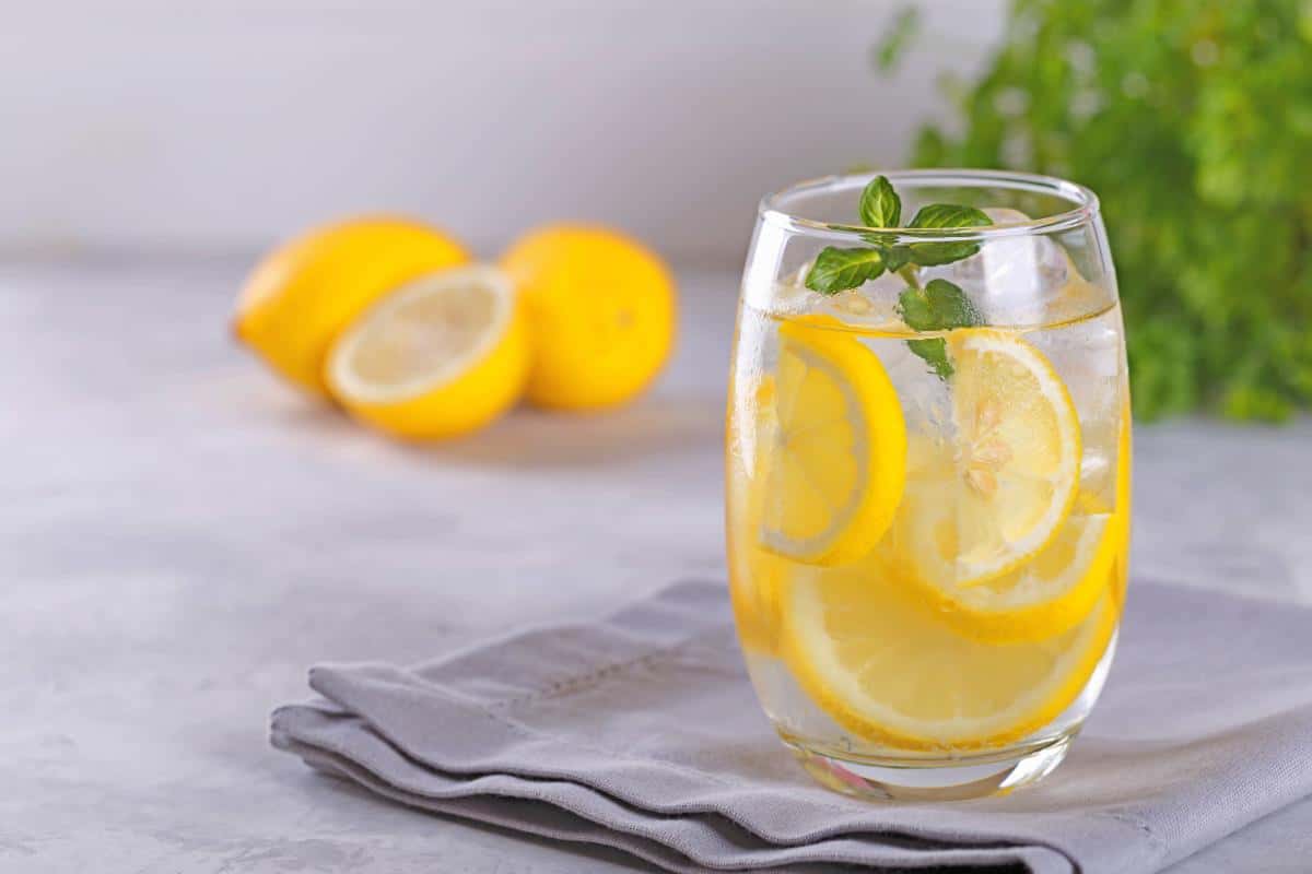 A glass of fruit infused water with lemon slices and mint leaves sits on a gray napkin, while two whole lemons and a lemon half add a refreshing touch in the blurred background—a perfect choice among healthy drinks featuring fresh fruits.