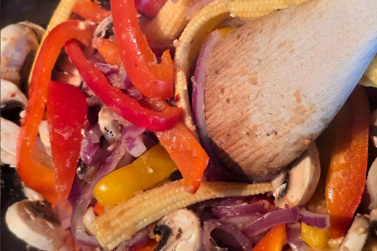A close-up of a wooden spoon stirring a colorful mix of sliced red and yellow bell peppers, baby corn, mushrooms, and red onions—perfect veggies for a Healthy Fakeaway or as a side to your favorite Sticky Beef Recipe.