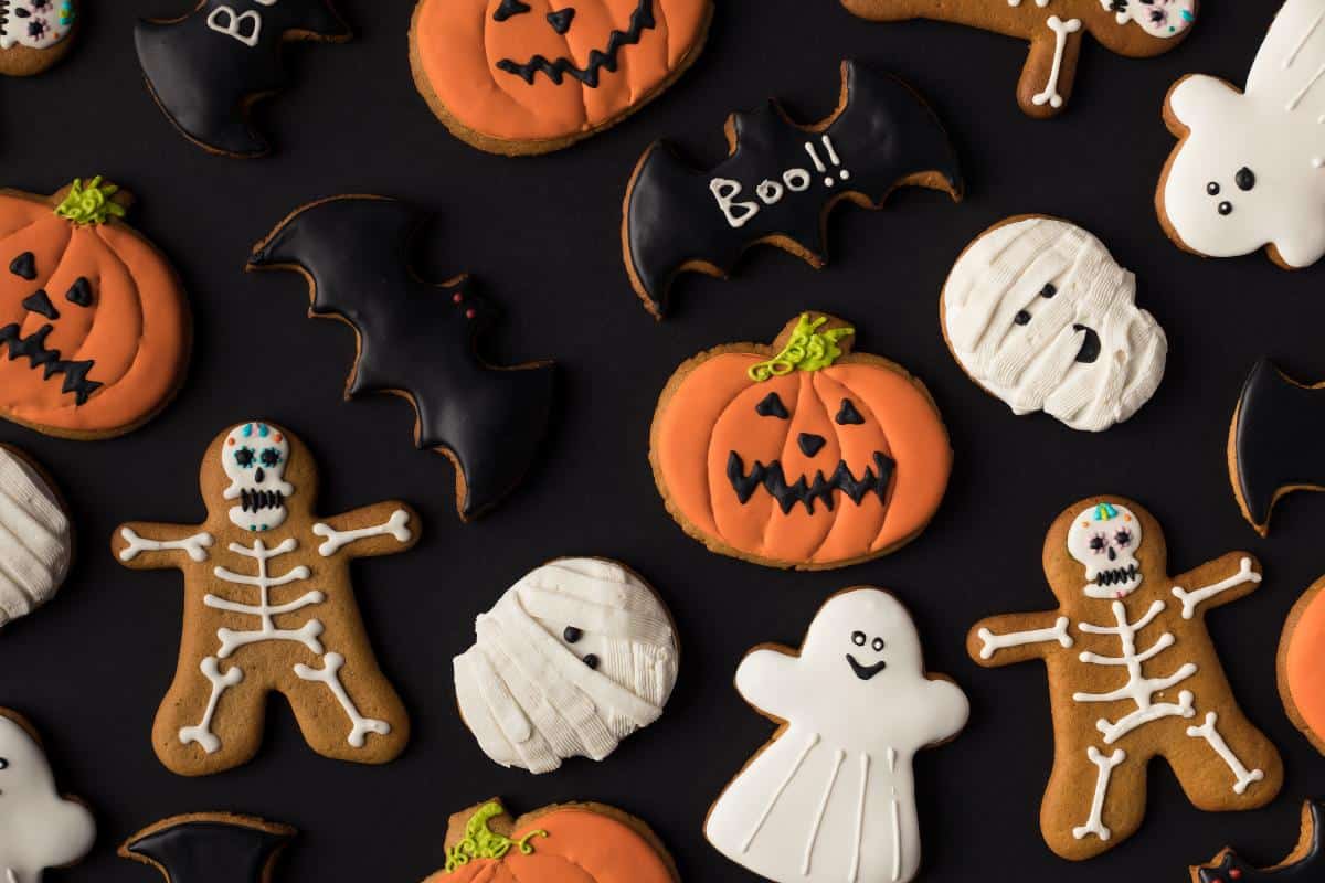 A variety of spooky treats—Halloween-themed cookies shaped like pumpkins, bats, ghosts, skeletons, and mummies—decorated with colorful icing are artfully arranged on a black background.