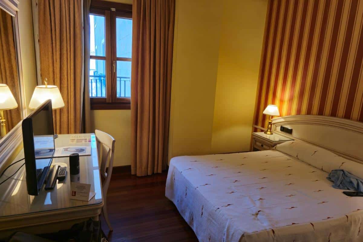 A cozy hotel room in one of the Best Hotel Chains features a double bed, striped wallpaper, bedside lamps, a desk with TV, chair, and a window with beige curtains letting in daylight—a perfect choice for Budget Hotels France seekers.