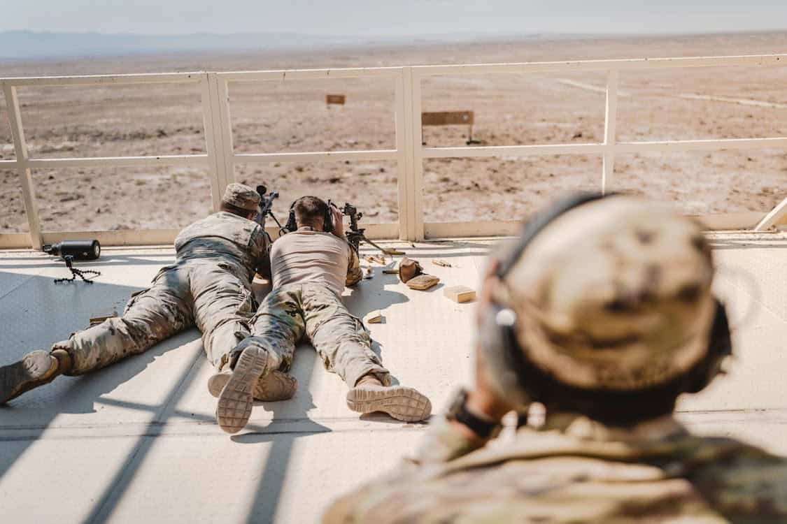 Three people in military uniforms are on a raised platform overlooking a desert landscape. Two are lying prone and aiming rifles at distant targets, while one is seated in the foreground observing.