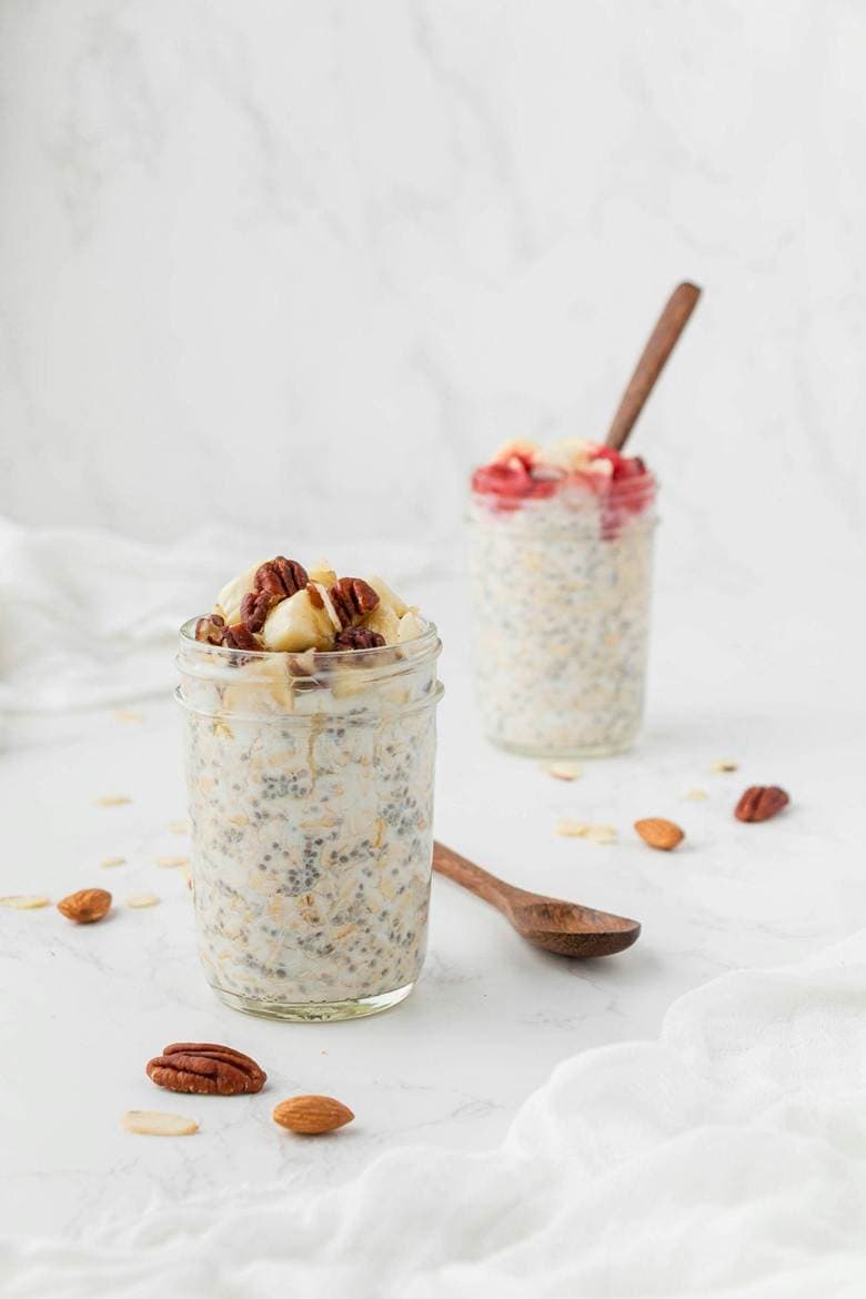 Two jars of overnight oats with whey protein topped with nuts and fruit sit on a white surface. One jar is garnished with pecans and banana slices; the other with strawberry pieces. Wooden spoons and scattered nuts are nearby.