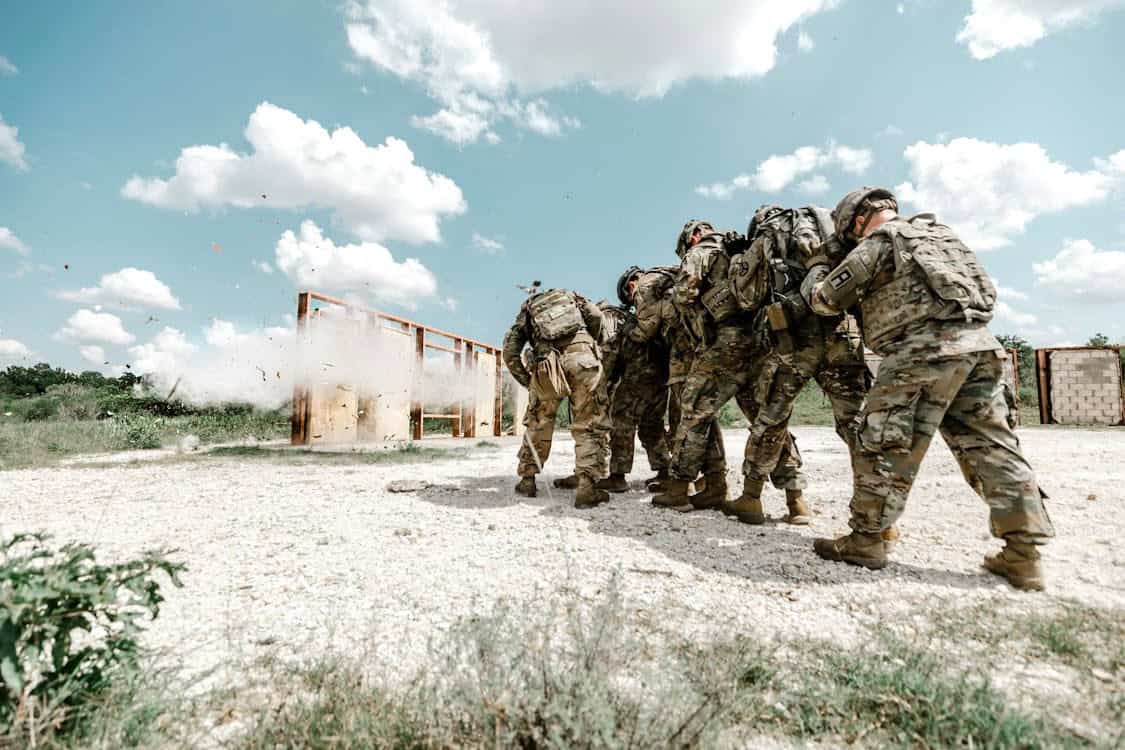 A group of soldiers in camouflage gear huddle together outdoors, moving forward as an explosion or blast occurs near a wooden structure in the background under a partly cloudy sky.