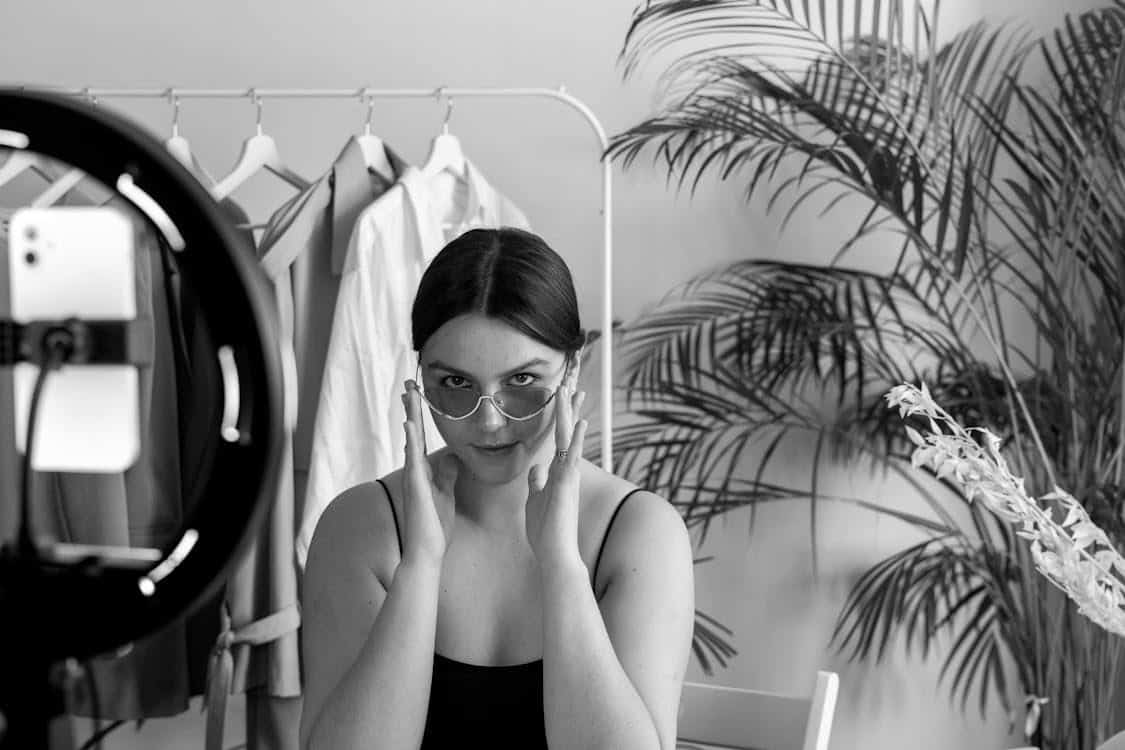 A woman adjusts her sunglasses and looks at a smartphone camera on a ring light stand, with clothes on a rack and potted plants in the background.