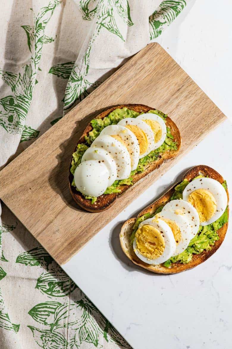 Two slices of toasted bread topped with mashed avocado and sliced hard-boiled eggs, sprinkled with black pepper, on a wooden board, with a patterned cloth nearby.