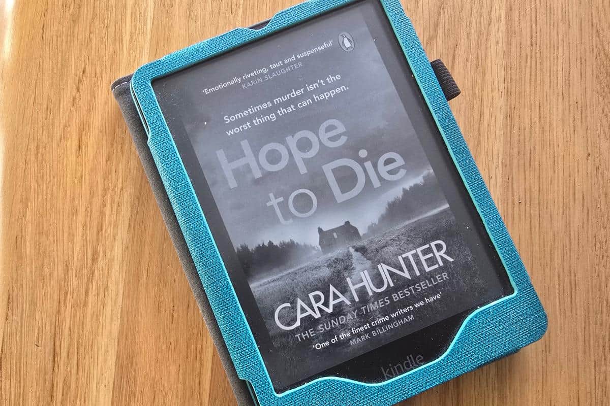 A Kindle in a blue case displays the cover of "Hope to Die" by Cara Hunter, showing a grayscale rural landscape with a house and trees, perfect for your Midweek Catch-Up, placed on a wooden surface.