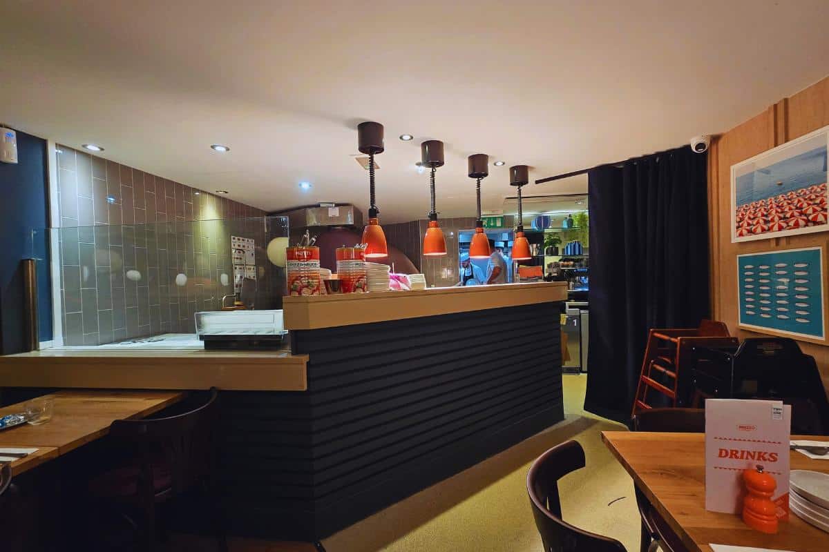 A cozy Prezzo Italian Nottingham interior features wooden tables, chairs, a service counter under red pendant lights, and shelves filled with items. A drinks menu sits on a table while framed artwork decorates the walls—perfect for your next restaurant review.