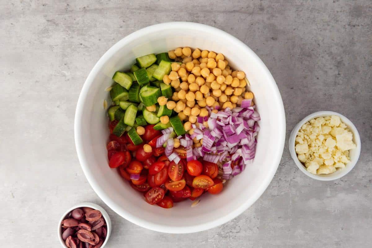 A white bowl with chopped cucumbers, cherry tomatoes, chickpeas, and red onions is arranged in sections for the ultimate healthy salad. Two smaller bowls hold sliced olives and crumbled feta cheese. The background is a gray surface.