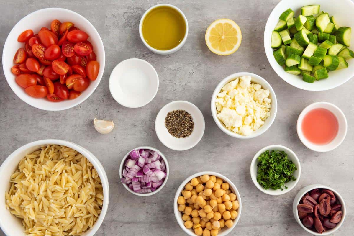 Bowls with Healthy Orzo Salad Recipe ingredients—grape tomatoes, cucumber, red onion, chickpeas, feta cheese, olives, parsley, olive oil, lemon, vinegar, and garlic—are artfully arranged on a gray surface.