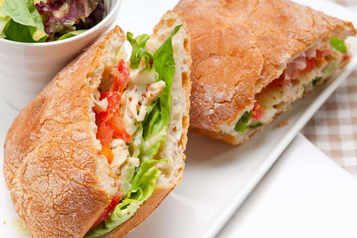 A close-up of a sandwich cut in half, filled with shredded chicken, lettuce, and tomato on crusty bread. Perfect inspiration for homemade paninis, with a small bowl of salad in the background on a white rectangular plate.