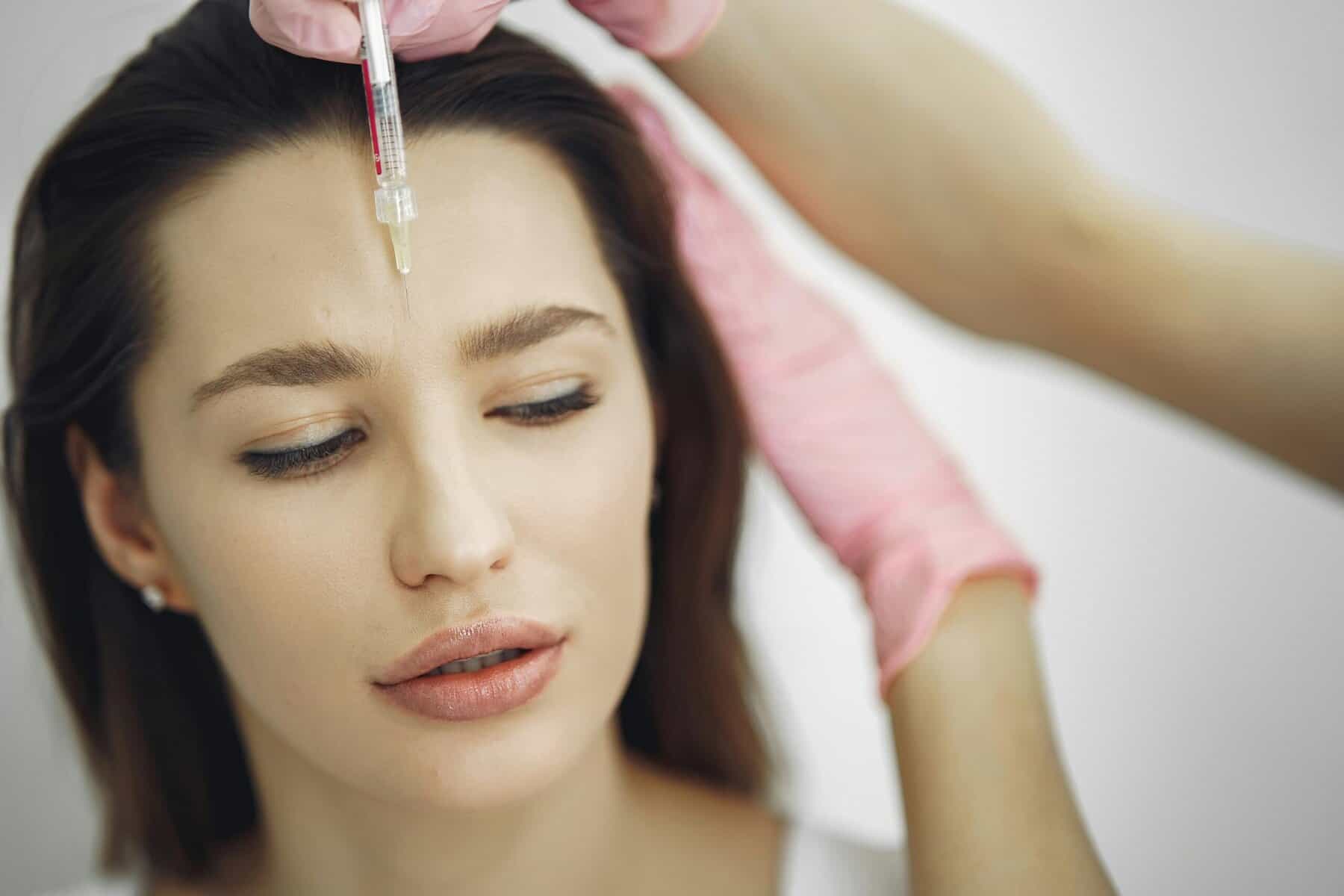 A woman with straight brown hair receives dermal fillers in her forehead from a professional wearing pink gloves. With a neutral expression and closed eyes, she embraces self-care routines for sculpted confidence.