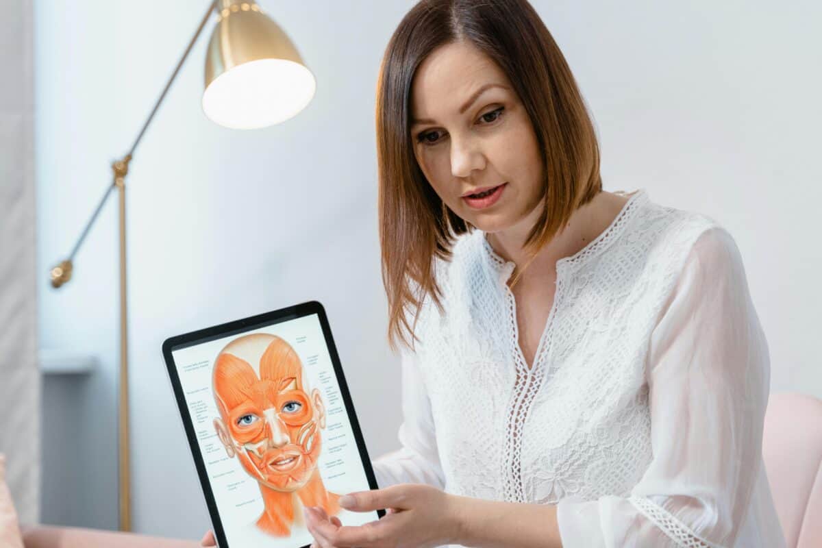 A woman in a white blouse holds a tablet displaying a detailed facial anatomy illustration, explaining the diagram—highlighting its importance for safer beauty treatments. A lamp and light furnishings are visible in the background.