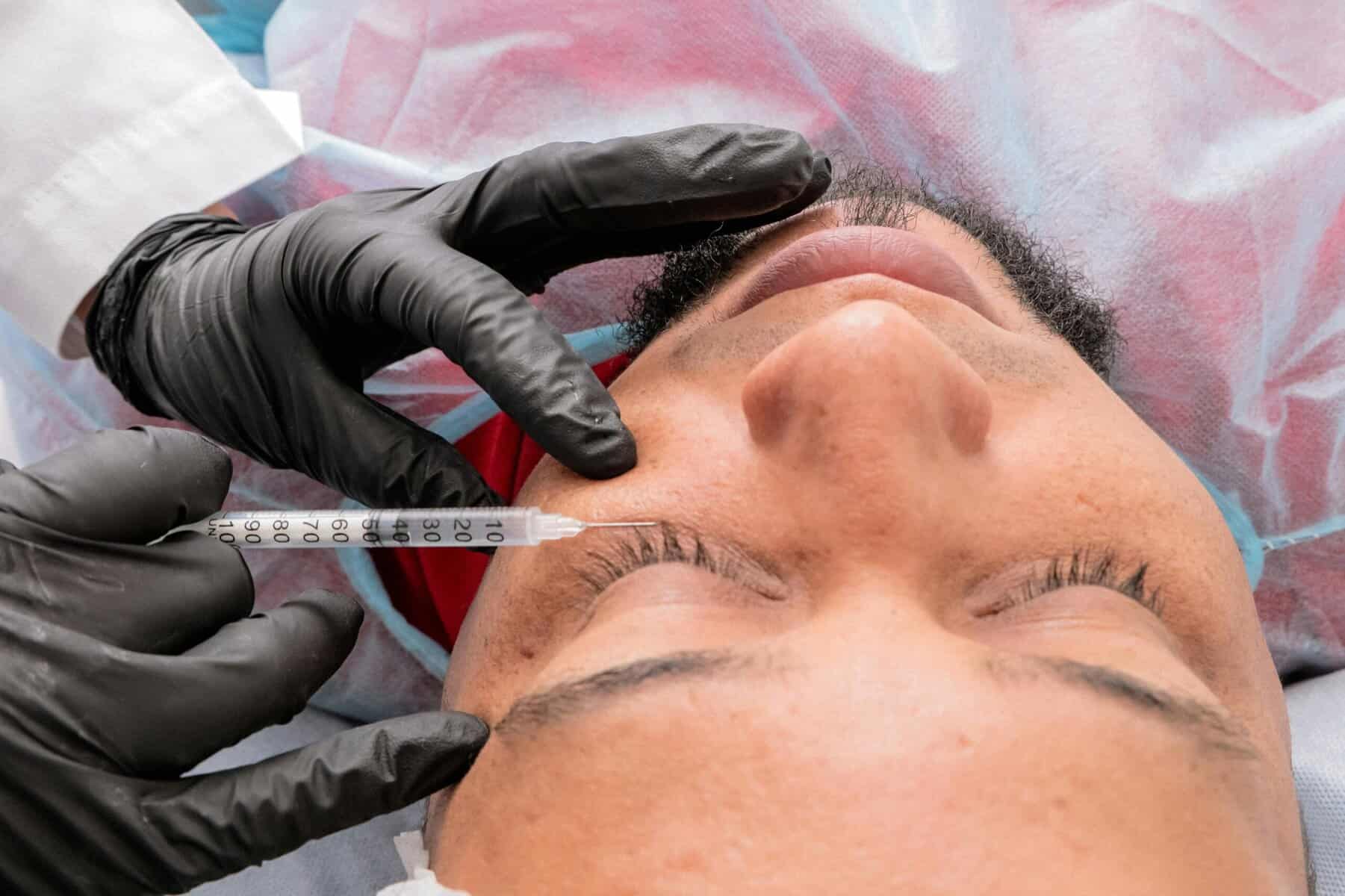 A person lying down with eyes closed receives a Restylane injection under the eye from a professional in black gloves and a lab coat, likely aiming for fuller, fresher natural features through a cosmetic or medical procedure.