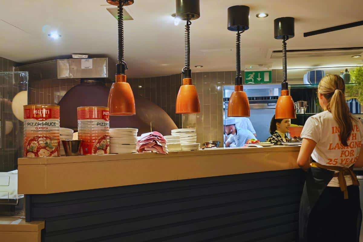 A woman stands at a Prezzo Italian Nottingham counter with orange hanging lights above. Stacks of plates, pizza sauce cans, and a wood-fired oven are visible, while chefs work in the open kitchen and another woman waits near the counter.
