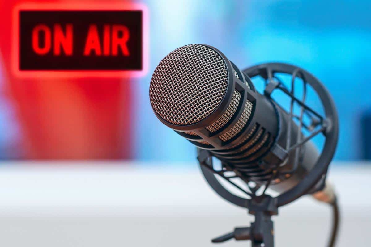 A close-up of a studio microphone on a stand with a blurred red "ON AIR" sign illuminated in the background, setting the stage for the Midweek Catch-Up live broadcast.