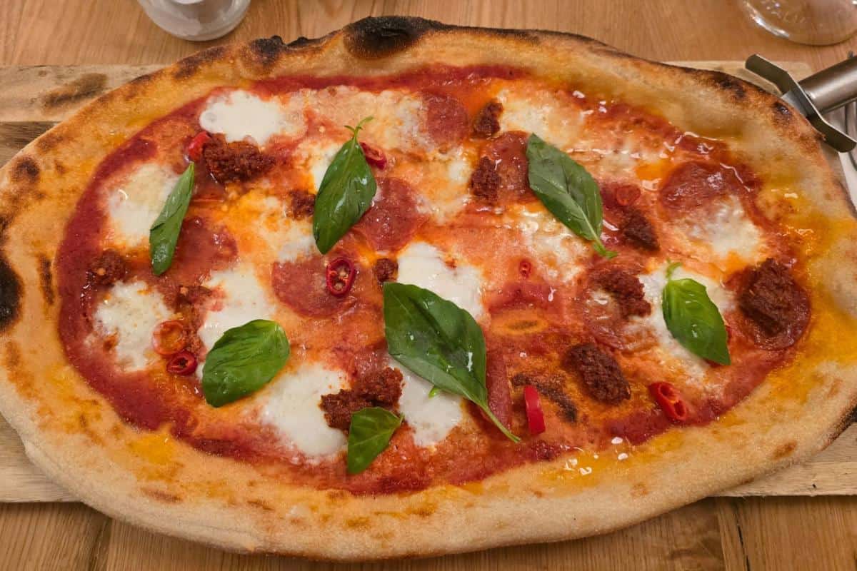 Oval-shaped pizza with a crispy, charred crust, topped with tomato sauce, melted cheese, pepperoni, red chili slices, spicy sausage dollops, and fresh basil leaves—just like you’d find at Prezzo Italian Nottingham.