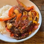A white bowl containing steamed white rice and Ultimate Sticky Beef stir-fried with red onions, baby corn, and red bell peppers, placed on a wooden surface—a perfect Healthy Fakeaway meal.