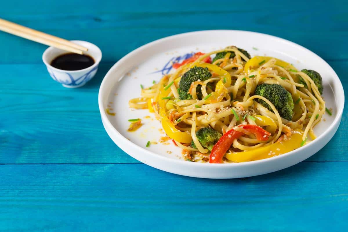 A plate of stir-fried noodles with broccoli and colorful peppers, topped with sesame seeds, sits on a blue wooden table. Enjoy this healthy takeaway alternative with chopsticks and a small dish of homemade stir-fry sauce.