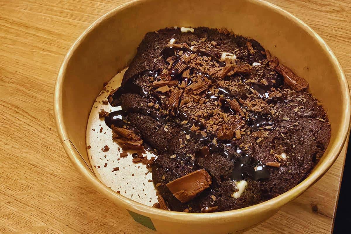 A round cardboard container holds a rich chocolate dessert topped with chocolate sauce, chocolate shavings, and small pieces of chocolate bars on a wooden surface—an indulgent treat reminiscent of the sweet offerings at Prezzo Italian Nottingham.