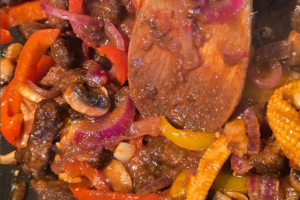 Close-up of a wooden spoon stirring a colorful mix of sautéed red and yellow bell peppers, red onions, mushrooms, and pieces of beef in a rich brown sauce—perfect for an Ultimate Sticky Beef healthy fakeaway at home.