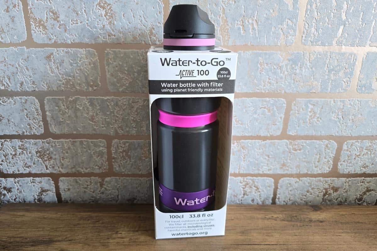 A Water-to-Go Active 100 water bottle with a filter, black with purple accents, stands upright in its packaging against a light brick wall and wooden surface. The packaging highlights features, capacity, and encourages you to stay hydrated throughout the day.