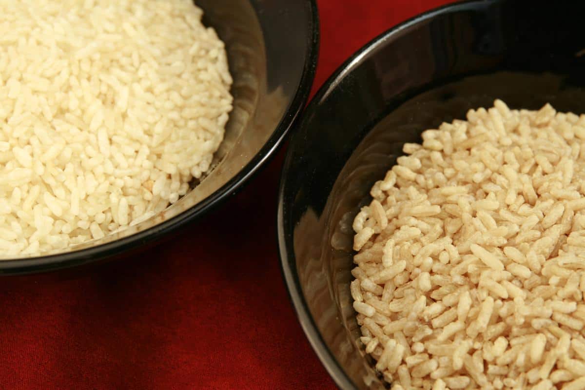 Two black bowls on a red surface highlight a white vs brown rice comparison: one holds cooked white rice, while the other contains brown rice.